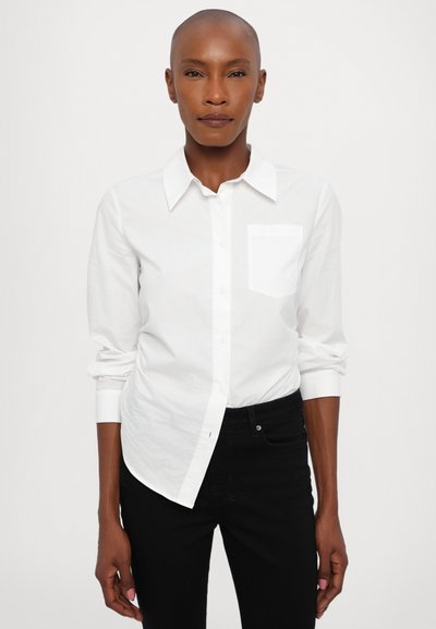 Camisa blanca de botones con cuello, un bolsillo en el pecho y mangas remangadas. Llevada con pantalones negros, de textura suave y corte ajustado.