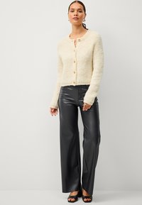 Gilet en crème pelucheux avec un col rond, fermeture à boutons sur le devant et manches longues, associé à un pantalon noir en simili cuir taille haute.