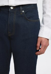 Primer plano de una persona que lleva vaqueros de mezclilla azul oscuro con costuras amarillas y una camisa blanca abotonada, mostrando la mano derecha relajada al lado.