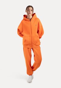 Orange zip-up hoodie med en stor lomme, parret med matchende sweatpants. Blødt stof, afslappet pasform og ribbede manchetter ved anklerne.