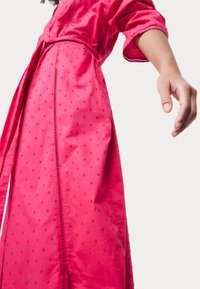 Abito rosa a un'altezza ginocchio con un motivo strutturato, vita con cintura e orlo dettagliato. Presenta maniche a tre quarti e sottili aperture lungo i bordi.
