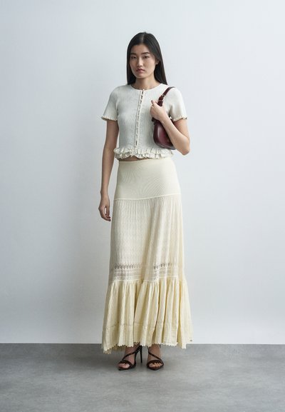 Νεαρή γυναίκα που φοράει μπεζ πλεκτό crop top και μακριά φούστα, κρατώντας μια μπορντό τσάντα ώμου και στέκεται σε γκρι πάτωμα μπροστά σε λευκό τοίχο.