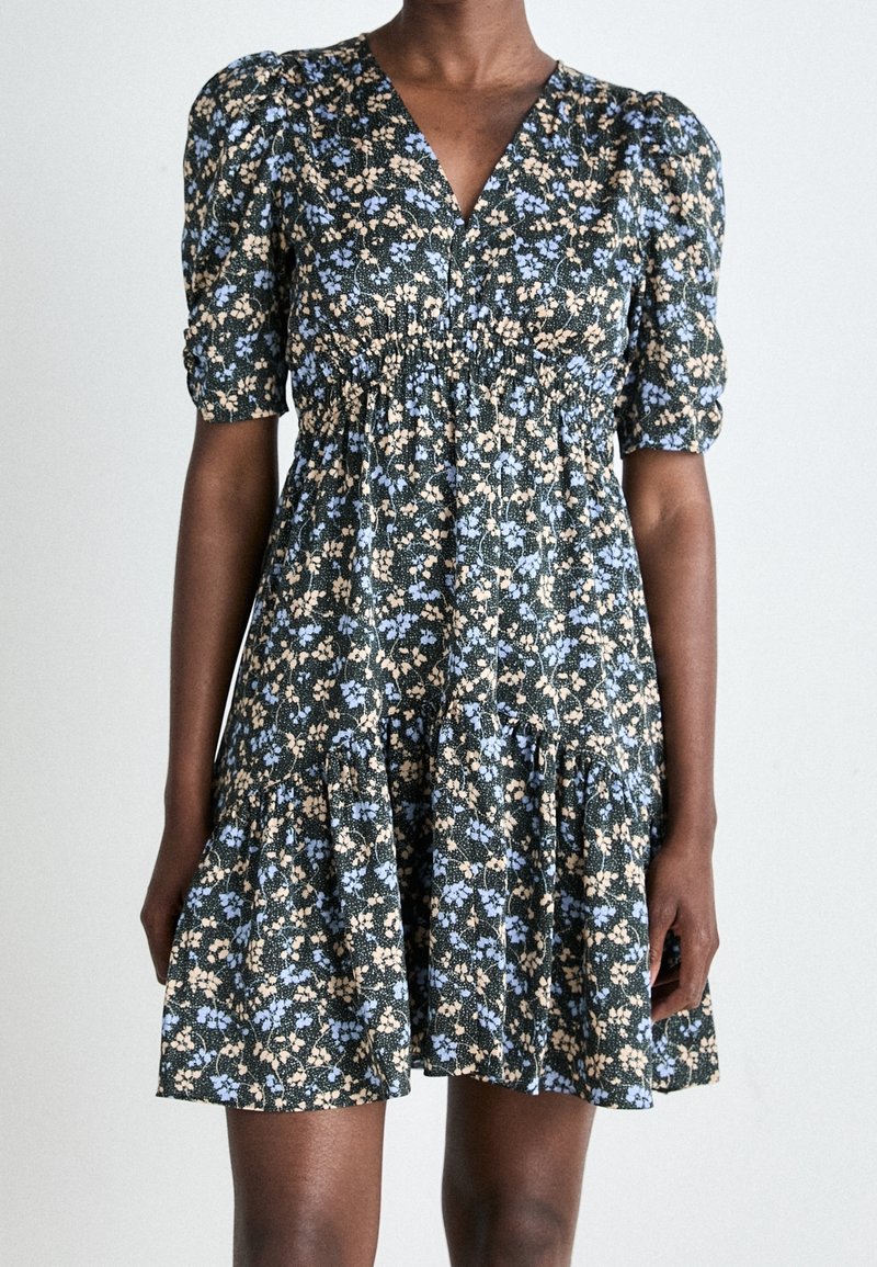 Femme portant une robe courte à manches courtes, col en V, longueur genoux, avec taille froncée et motif floral bleu et beige sur fond foncé.