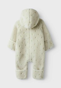 Krämfärgad fleeceoverall med en texturerad, dimplad mönstring, huva och uppvikta ärmslut. Mjuk, varm och designad för komfort.