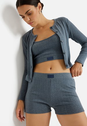 Femme portant un crop top côtelé gris assorti, un cardigan et un short, posant avec une main sur la hanche devant un fond blanc.