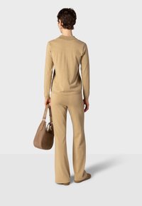 Personne aux cheveux courts portant un haut beige à manches longues et un pantalon, tenant un sac à main marron, debout de dos sur un fond uni.