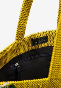 Żółta torba tote o plecionej strukturze z teksturowanym wykończeniem, wyposażona w czarne wnętrze z materiału i kieszonkę na zamek do bezpiecznego przechowywania.