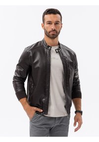 Giacca bomber in pelle marrone con zip frontale, polsini a coste e colletto alla cinese, indossata sopra una camicia grigio chiaro. Texture liscia e design aderente.