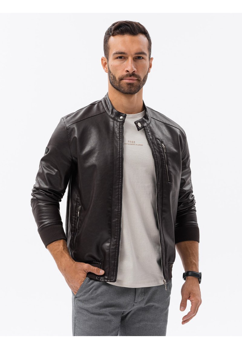 Giacca bomber in pelle marrone con zip frontale, polsini a coste e colletto alla cinese, indossata sopra una camicia grigio chiaro. Texture liscia e design aderente.