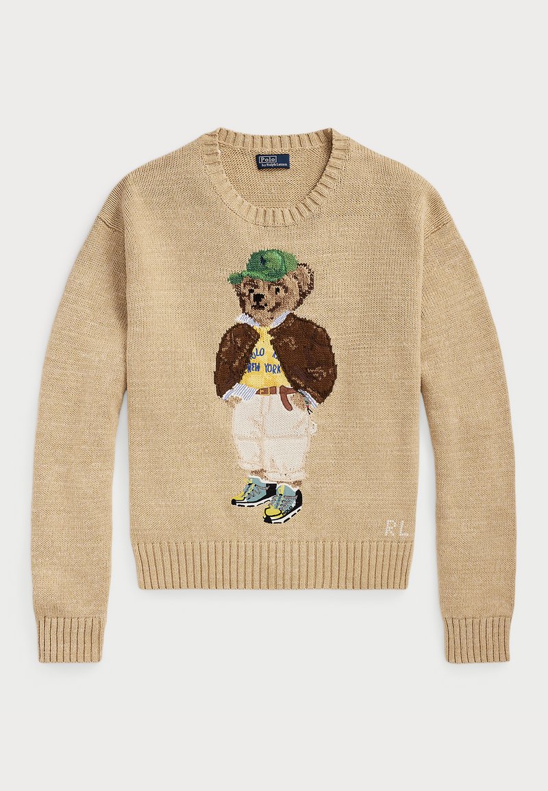 Suéter de punto beige con un personaje de oso bordado que lleva un sombrero verde, una camiseta amarilla, una chaqueta marrón y pantalones claros con zapatillas.