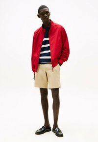 Veste bomber rouge sur une chemise à rayures navy, associée à un short beige et des mocassins noirs. Textures lisses, design décontracté et couleurs vibrantes.