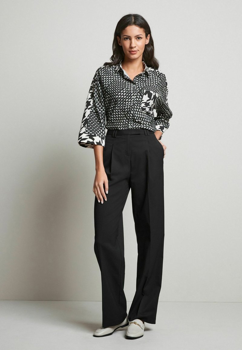 Pantalon noir taille haute à jambe large, associé à un blouse à motifs noir et blanc avec des manches évasées et une fermeture à boutons.
