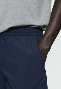 Pantaloni corti blu navy con cintura elastica e tasche laterali, realizzati in materiale leggero, abbinati a una maglietta grigia.