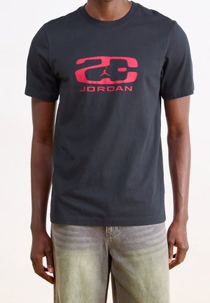 Mand iført sort t-shirt med rød "23 Jordan" logo og grå falmede jeans mod en ensfarvet baggrund.
