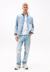 Hellblauer Denim-Jacke mit einer aufgesetzten Tasche, kombiniert mit hellblauen, ausgewaschenen Jeans. Model trägt ein weißes T-Shirt und mehrfarbige Sneakers.