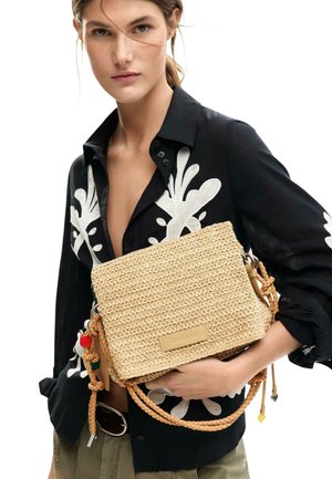 Mujer con camisa negra bordada en blanco sostiene un bolso tejido beige con asas trenzadas y cuentas decorativas.