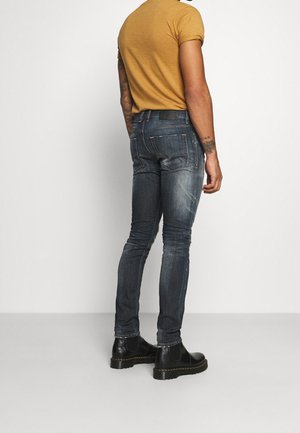 Homme portant un jean slim bleu délavé, une chemise jaune moutarde à manches courtes, des bottes en cuir noir, debout devant un fond blanc.