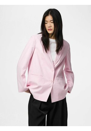 Kvinde med langt sort hår iført en lyserød blazer over en hvid top og løse sorte bukser, stående med hænderne i lommerne.