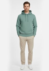 Sudadera con capucha verde texturizada con cordón ajustable, combinada con pantalones beige y zapatillas blancas. Diseño simple con un ajuste casual.