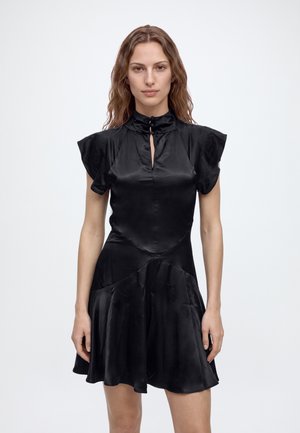 Jeune femme aux cheveux ondulés porte une courte robe en satin noire avec un col montant et des manches courtes, debout devant un fond clair uni.