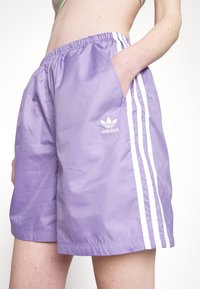 Pantaloncini sportivi viola realizzati in tessuto leggero, con logo Adidas bianco, cintura elastica e strisce laterali bianche.
