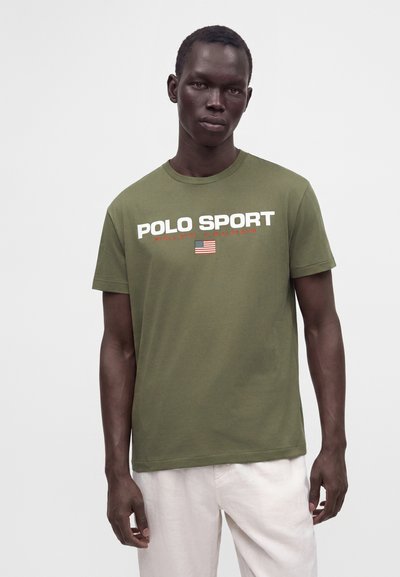 Νέος άντρας που φοράει ένα ελιάς πράσινο T-shirt Polo Sport με τη σημαία των ΗΠΑ και λευκό παντελόνι σε απλό φωτεινό φόντο.