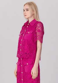 Haut en dentelle fuchsia à manches courtes, avec un col, des poches avant et des boutons dorés. Motif floral distinct sur tout le tissu.