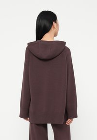 Mulher com cabelo preto comprido usa um hoodie de malha solto em mauve escuro e calças combinando, de costas para o espetador, em frente a um fundo branco liso.