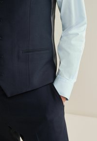 Person wearing a dark navy vest and pants with a light blue long-sleeve shirt, hand casually in pocket against a neutral background.