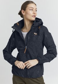 Giacca impermeabile navy con pois bianchi, cappuccio regolabile, zip frontale e dettagli in pelle marrone. Tessuto morbido e polsini elastici.