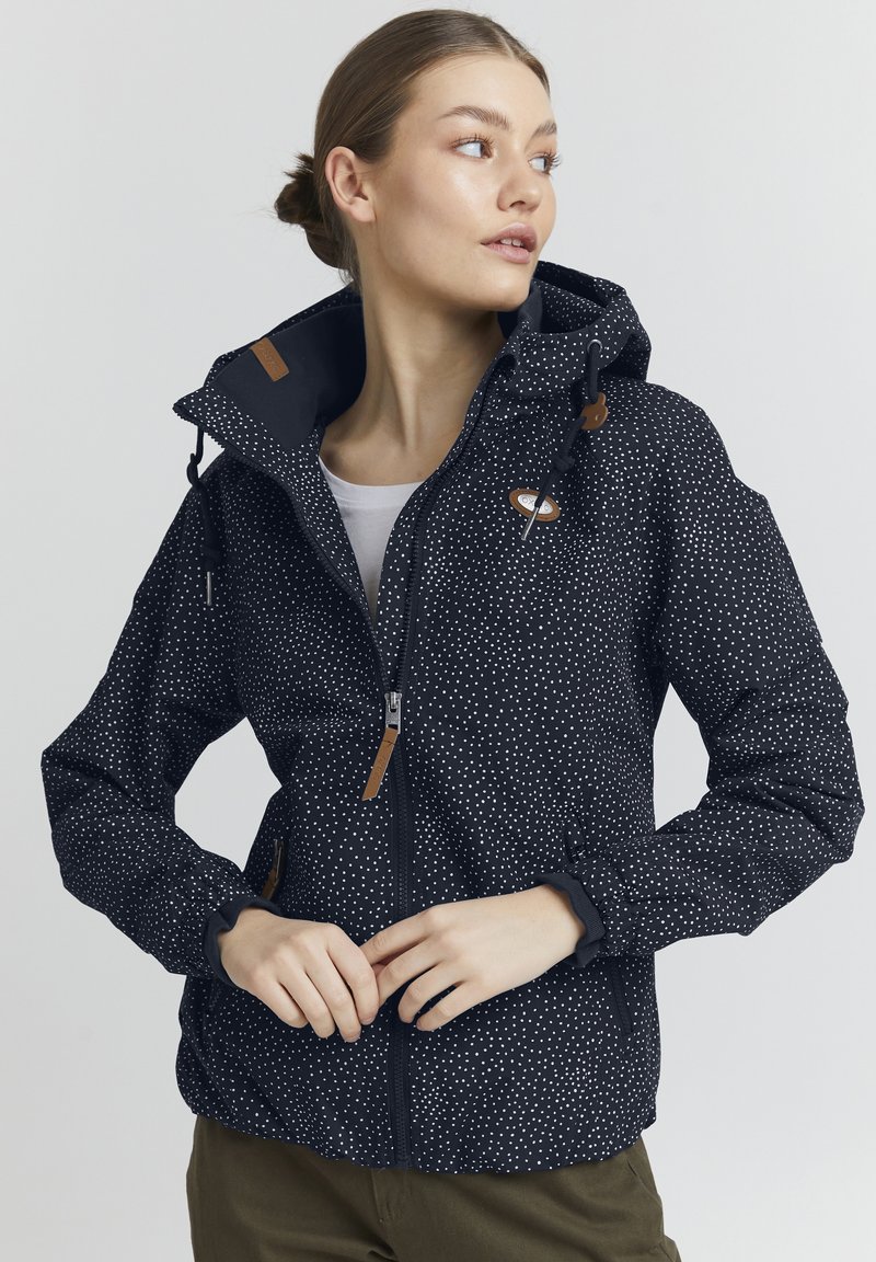 Giacca impermeabile navy con pois bianchi, cappuccio regolabile, zip frontale e dettagli in pelle marrone. Tessuto morbido e polsini elastici.