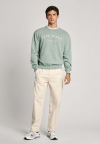 Gaiši zaļš sporta krekls ar uzrakstu "Pepe Jeans London W11", drukātu baltā krāsā, saskaņots ar krēmkrāsas biksēm un baltām sporta apaviem.