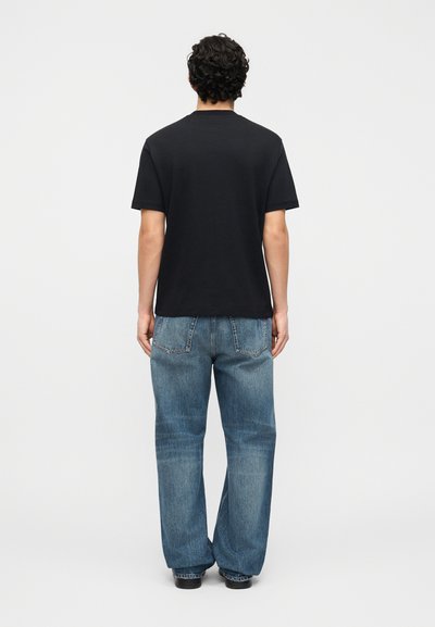 Άνδρας με σγουρά σκοτεινά μαλλιά στέκεται με την πλάτη, φοράει μαύρο κοντομάνικο μπλουζάκι, άνετα μπλε τζιν και μαύρα παπούτσια σε λευκό φόντο.