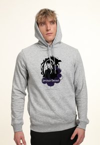 Giovane con capelli biondi scompigliati indossa un hoodie grigio con una grafica nera e viola che raffigura una donna, un corvo e il testo "Socialmente Distanziati."