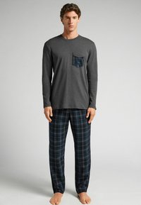 Camiseta de manga larga en gris carbónico con un bolsillo de cuadros, combinada con pantalones de pijama a cuadros en tonos azul oscuro, verde y blanco. Descalzo.