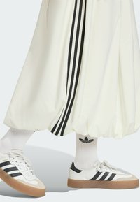 Bijele balon hlače s crnim bočnim trakama, uparene s bijelim Adidas Samba tenisicama s crnim naglascima i gumiranim đonom, te bijelim čarapama do gležnja.