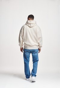 Beige hooded sweatshirt with oversized fit, cuffed sleeves, and kangaroo pocket, paired with light blue denim jeans and white sneakers.