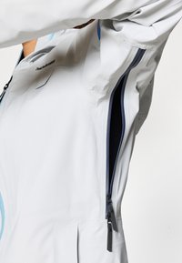 Vaaleanharmaa takki, jossa on tyylikäs muotoilu, vetoketjulla varustettu avaus ja kontrastiväriset laivastonsiniset yksityiskohdat. Liukas pinta ja istuva siluetti.