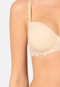 Beige rintaliivi, jossa on muotoillut kupit, pitsiysitykset ala reunassa ja ohuet säädettävät olkaimet. Sujuva pinta, jossa on hienovarainen kiilto.