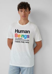 Junger Mann, der an einer weißen Wand steht, trägt ein weißes T-Shirt mit "Human Beings" in Regenbogenfarben und dem Text "Farben können variieren".