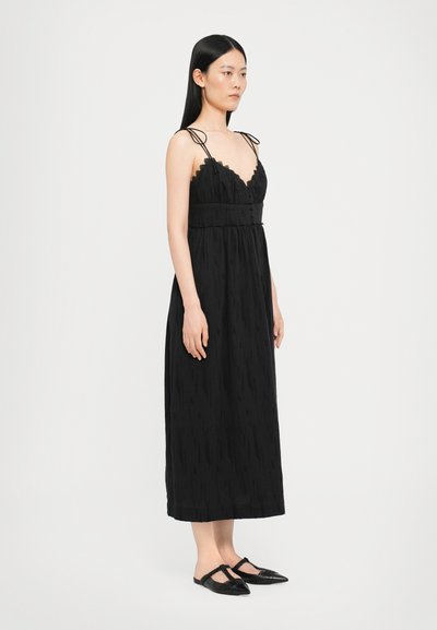Schwarzes Maxikleid mit Spaghetti-Trägern, strukturiertem Stoff und angepasstem Oberteil. Verfügt über eine Spitzenborte und eine taillierte Taille. Ein Paar schwarze Ballerinas.