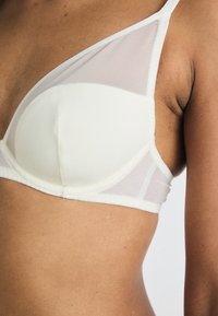 Bha met een witte, gevormde cup met doorzichtige meshpanelen en verstelbare bandjes; gladde textuur met minimale naden en zachte randen.