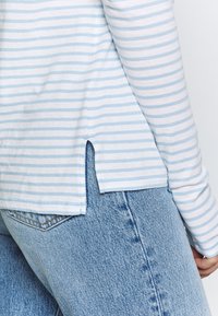 Camicetta a maniche lunghe a righe blu chiaro e bianco con spacchi laterali, abbinata a jeans in denim blu chiaro. Il tessuto sembra morbido e traspirante.