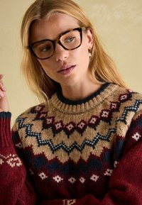 Strickpullover mit mehrfarbigen geometrischen Mustern in Beige, Burgunderrot und Marineblau. Mit geripptem Halsausschnitt und strukturierten Ärmeln. Klare Brille.