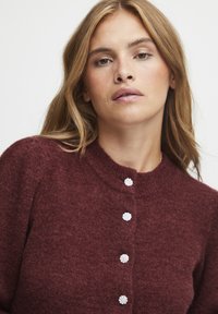 Bordeauxfarbener Strickcardigan mit rundem Ausschnitt, der vorne mit perlenähnlichen Knöpfen versehen ist. Weiche Textur und taillierte Passform.