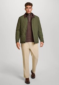 Quilted olive green jacket with a brown zip-up sweater underneath, beige trousers, and dark brown shoes. Casual design, comfortable fit.