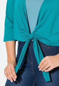 Top court teal avec un nœud à l'avant, porté avec une chemise bleu clair en dessous. Jean en denim foncé avec des détails de couture visibles.