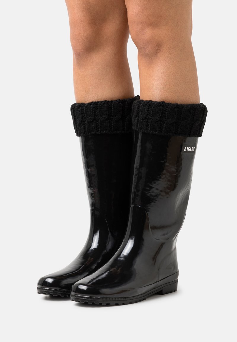 Bottes de pluie en caoutchouc noir avec une finition brillante et des poignets en tricot noir côtelé. Dotées d'une semelle texturée pour une meilleure adhérence. Logo de la marque visible.