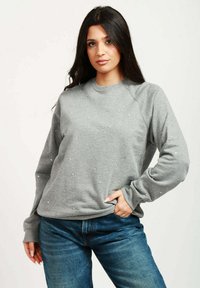 Felpa grigia con scollo a girocollo, decorata con piccole borchie, presentante maniche raglan e una vestibilità morbida. Indossata con jeans blu.