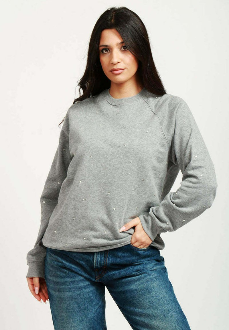 Felpa grigia con scollo a girocollo, decorata con piccole borchie, presentante maniche raglan e una vestibilità morbida. Indossata con jeans blu.
